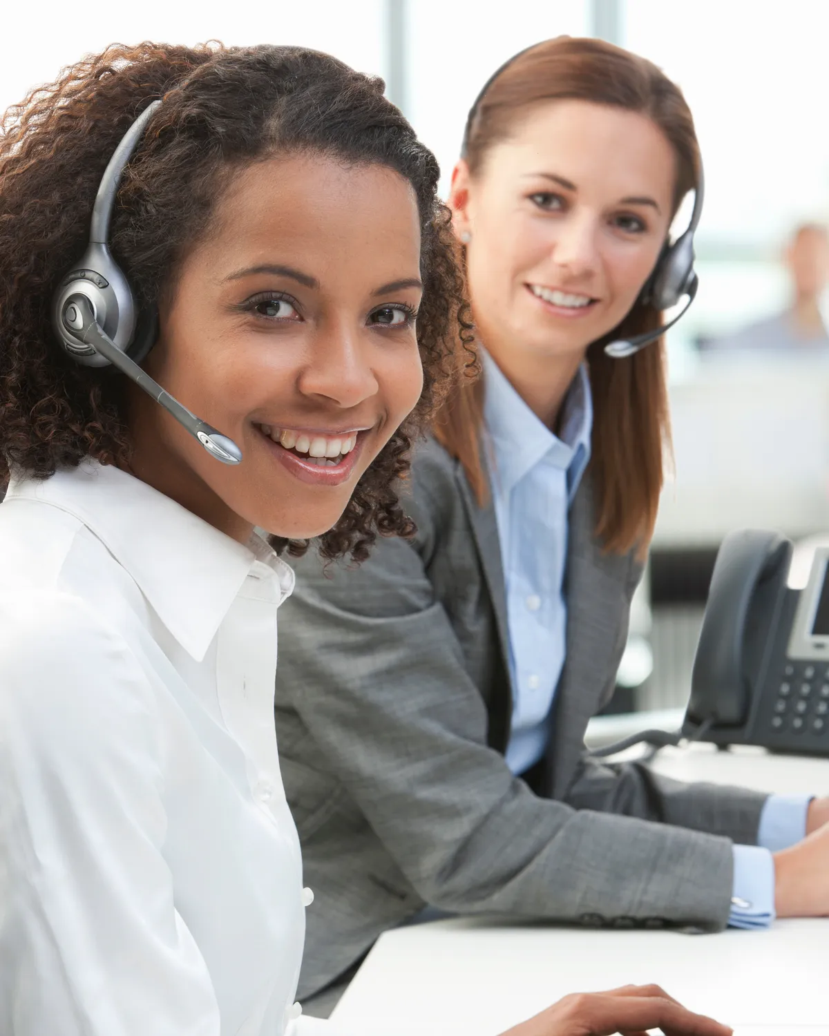 Two front-desk staff in headsets smiling at their desks, an IP desk phone beside them.