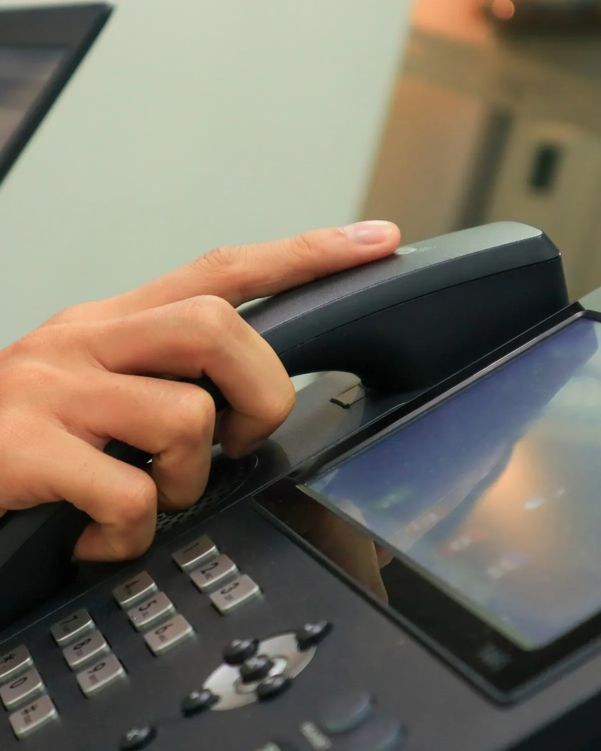A hand lifting the handset of a modern IP desk phone with a color touchscreen.